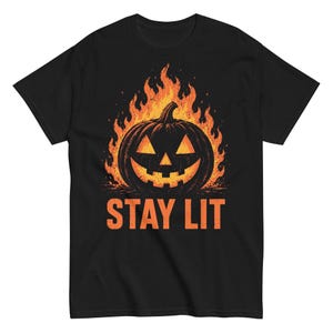 Puede incluir: Una camiseta negra de manga corta con un gráfico de una calabaza de Halloween rodeada de llamas naranjas y amarillas. Debajo de la calabaza, el texto STAY LIT está impreso en letras mayúsculas de color naranja, centrado en el pecho.