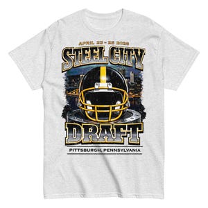 Puede incluir: Camiseta gris claro con un diseño de casco de fútbol americano negro y amarillo. El diseño incluye las palabras "Steel City Draft", las fechas "25-26 de abril de 2026" y "Pittsburgh, Pensilvania".