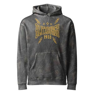 May include: A charcoal grey hoodie with a distressed, mottled texture featuring a yellow graphic on the chest. The design includes the text BLITZBURGH above the year 1933, surrounded by four small star icons and four lightning bolt symbols.