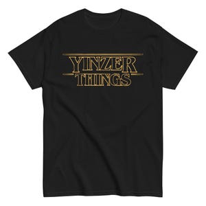 以下が含まれることがあります： 黒いTシャツ。前面に「Yinzer Things」という文字が金色の様式化されたフォントでプリントされています。テキストは中央に配置されています。Tシャツは半袖で、クルーネックです。