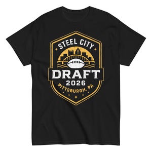 May include: Black t-shirt featuring a gold and white graphic. The graphic includes the text "Steel City Draft 2026 Pittsburgh, PA" with an image of a bridge and a football.