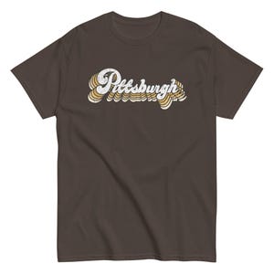 May include: A brown t-shirt with the word "Pittsburgh" in a retro font. The text is white with yellow and cream-colored accents, creating a layered effect. The shirt has short sleeves and a crew neck.