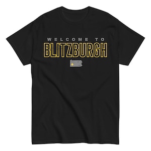 May include: Black t-shirt with the text "WELCOME TO BLITZBURGH" in yellow and gray lettering. A gray outline of Pennsylvania with a yellow star is below the text. The shirt is a short-sleeved crew neck.