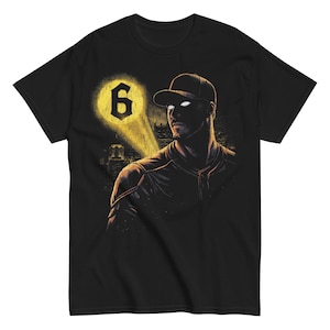 Puede incluir: Camiseta negra con una ilustración estilizada de un jugador de béisbol con ojos brillantes y una gorra de béisbol. Un foco amarillo ilumina el número 6. El fondo muestra un horizonte urbano.