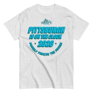 Puede incluir: Camiseta gris claro con el texto "PITTSBURGH IS ON THE CLOCK 2026" en turquesa y blanco. La camiseta también presenta el texto "PROUDLY FORGING THE FUTURE" en un diseño curvo. Un logotipo está en la parte superior.