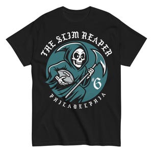 May include: Black t-shirt featuring a cartoon grim reaper holding a football and a scythe. The design includes the text "THE SLIM REAPER" and "PHILADELPHIA" in white, with a teal and white circular background. The number "6" is also included.