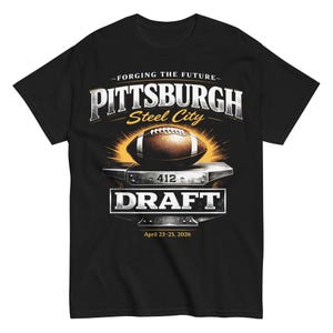 Könnte beinhalten: Schwarzes T-Shirt mit einem Grafikdesign, das einen Football, einen Amboss und den Text "PITTSBURGH Steel City DRAFT" mit dem Datum "23.-25. April 2026" zeigt. Das Design beinhaltet einen Sonnenstrahleffekt hinter dem Football.