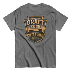 Puede incluir: Camiseta gris con un gráfico dorado y negro. El gráfico dice "DRAFT 2026 PITTSBURGH" con un diseño de fútbol americano. La camiseta es de cuello redondo y manga corta.