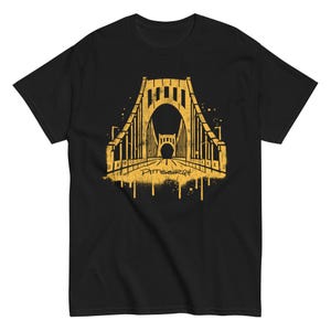 以下が含まれることがあります： 黒いTシャツ。金色の橋のグラフィックと、下に「Pittsburgh」の文字。橋のデザインは、スプレーペイントのようなスタイルです。シャツは柔らかい素材で作られています。