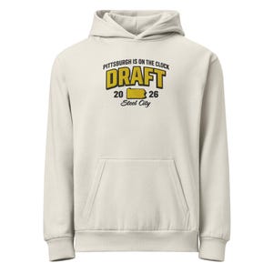 Puede incluir: Sudadera con capucha color crema con bolsillo delantero y capucha con cordón. La parte delantera presenta el texto "PITTSBURGH IS ON THE CLOCK DRAFT 20 26 Steel City" en amarillo y negro. Una sudadera casual y cómoda.