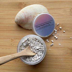 May include: A clear glass jar filled with white crushed coral, a wooden spoon, and a smaller jar with a label that reads "Penny Pincher Food Co. Your one stop hermit shop!"