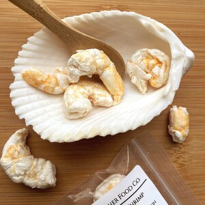 May include: Dehydrated shrimp with orange and white coloring, presented in a white seashell with a wooden spoon. A small bag of shrimp is visible in the bottom right corner. The shrimp are on a wooden surface.