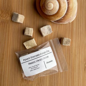 May include: A clear plastic bag containing freeze-dried duck breast pieces, with a label that reads "PENNY PINCHER FOOD CO. FREEZE DRIED DUCK." The label also states "100% Duck Breast, 70% Protein, 100% Delicious!" A seashell is also visible.
