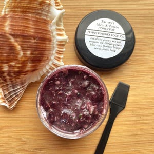 May include: An overhead shot features a jar of Barney's Meat & Potato Honey Pot, a black lid with text, a black spatula, and a seashell. The jar contains a dark purple mixture. The lid reads "PENNY PINCHER FOOD CO."