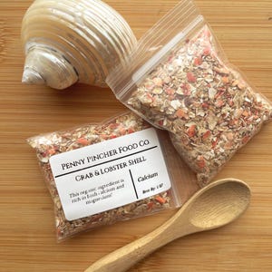 May include: A seashell, two small clear bags filled with crab and lobster shell flakes, and a wooden spoon rest on a wooden surface. The bags have labels that read "PENNY PINCHER FOOD CO. CRAB & LOBSTER SHELL". The text also states the product is rich in calcium and magnesium.