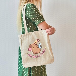 May include: A beige tote bag with a cartoon illustration of a capybara wearing a party hat and fairy wings. The text "She was a fairy!" is printed on the bag.