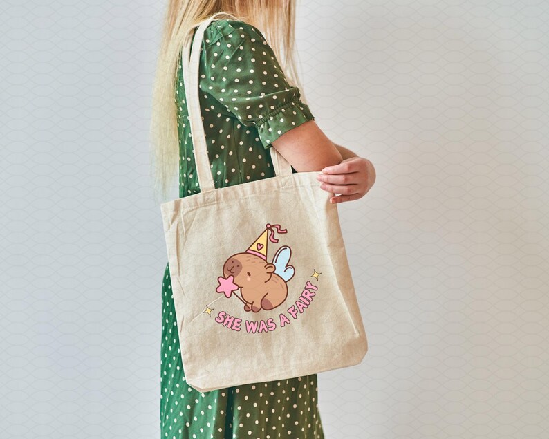 May include: A beige tote bag with a cartoon illustration of a capybara wearing a party hat and fairy wings. The text "She was a fairy!" is printed on the bag.
