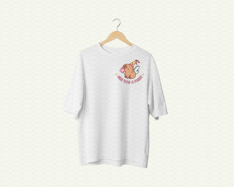 May include: White t-shirt with a cartoon illustration of a brown animal wearing a party hat and fairy wings. The text "See you're a fairy" is printed below the illustration.