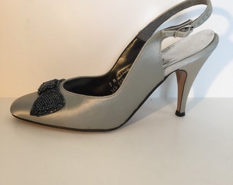 Puede incluir: Un par de tacones de slingback de satén gris claro con un lazo negro de cuentas en la punta. Los tacones miden aproximadamente 7,5 cm de alto.