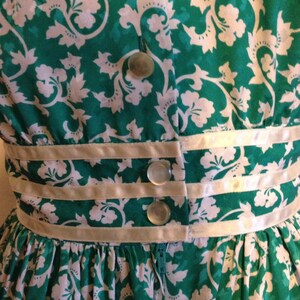 May include: A green and white floral dress with a white satin waistband and three white buttons. The dress has a zipper in the back.