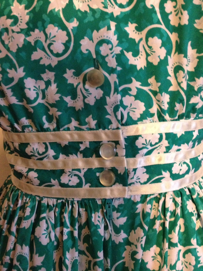 May include: A green and white floral dress with a white satin waistband and three white buttons. The dress has a zipper in the back.