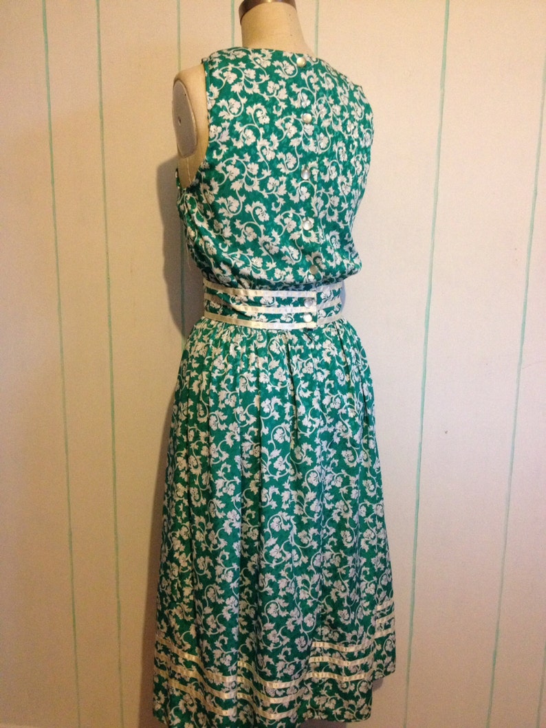 May include: A green and white floral print dress with a white ribbon belt and white trim around the bottom hem.