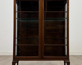 May include: A dark wood display cabinet with two glass doors and glass shelves. The cabinet has a decorative carved top and stands on curved legs. The interior is dark, and the glass doors offer a view of the shelves.