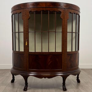 May include: A mahogany display cabinet with a curved front and glass panels. The cabinet features a keyhole with a gold tassel and stands on four curved legs. The interior is a light colour, and the wood grain is visible.