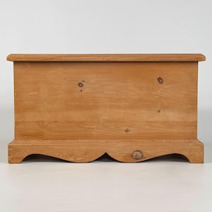 May include: A light brown wooden chest with a rectangular shape. The chest has a flat top and a decorative, scalloped base. The wood grain is visible, and there are several knots in the wood. The chest is likely used for storage.