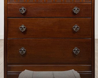 Puede incluir: Una cómoda de madera de color marrón oscuro con tres cajones horizontales y tiradores de metal oscuro ornamentados. El cajón superior presenta un patrón de arco tallado en el borde superior y la base se apoya sobre cuatro patas robustas.
