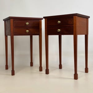 May include: A pair of matching wooden bedside tables, each featuring two drawers with small, round, polished brass knobs. The tables are constructed from a warm-toned wood with a smooth finish, standing on four slender, straight legs.