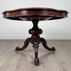 May include: A dark brown, antique, round table with a decorative pedestal base and three curved legs. The table has a scalloped edge and is made of wood. The table is approximately 76 cm in diameter.