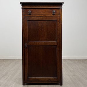 May include: A tall, dark brown wooden cabinet featuring a rectangular door with a central panel, a top drawer with two small metal knobs, and a single metal latch on the left side of the door. The wood grain is visible throughout the structure.
