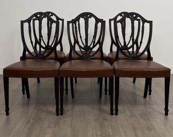 May include: A set of six antique dining chairs. The chairs have dark brown wooden frames with shield-shaped backs and brown leather seats. The seats are rectangular and have a studded trim. The chairs are arranged in a row.