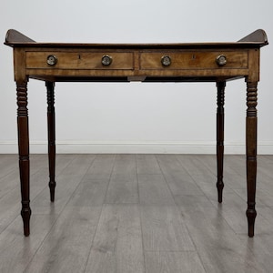 May include: A rectangular wooden desk featuring a flat top with raised side edges, two horizontal drawers with circular brass knobs, and four turned legs. The wood displays a natural grain pattern in a medium brown tone with a smooth finish.