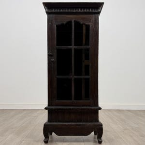 May include: A dark brown wooden cabinet with a glass door, featuring six panes. The cabinet has a decorative top and sits on a base with curved legs. The wood has a distressed finish, giving it a vintage appearance.