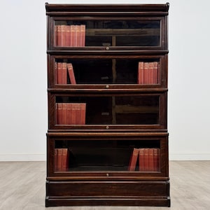 May include: A dark oak barrister bookcase with four glazed sections. Each section is filled with a row of red-covered books. The bookcase features a traditional design, with a curved top and a decorative base moulding.