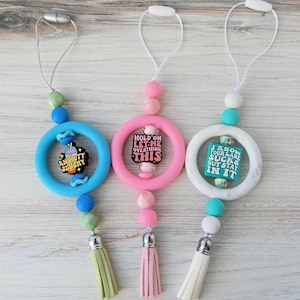 May include: Three silicone teething toys with different colored beads and tassels. The first toy has a blue ring with a blue and pink cartoon drink with the text "Anxiety Slushie". The second toy has a pink ring with a black square with the text "Hold on Let me Overthink This". The third toy has a white ring with a turquoise square with the text "I know your lane sucks but stay in it".