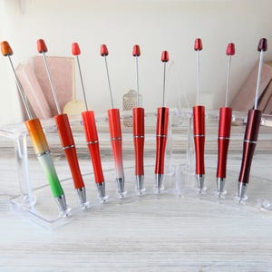 May include: A collection of nine red and silver pens with varying shades of red and orange, displayed in a clear acrylic stand. Each pen has a silver shaft and a small, rounded top. The pens are arranged in a row.