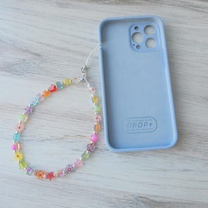 May include: A light blue phone case with a clear beaded phone strap. The phone case has the text "DROP+" printed on the back. The beaded strap is made of colorful glass beads in various shapes, including stars and flowers.