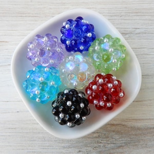 May include: A collection of colorful, spherical beads in a white dish. The beads are various colors, including blue, green, red, black, and clear, with small white accents. The beads have a clustered, textured appearance.