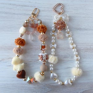 May include: Two beaded keychains with charms. One keychain is white with pearl beads and a heart charm. The other keychain is brown and white with pom poms and a heart charm.