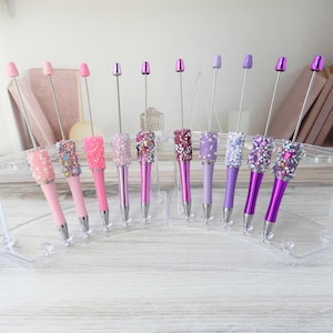 May include: A collection of decorative pens in various shades of pink and purple. Each pen features a bedazzled barrel and a matching colored top. The pens are displayed in a clear, angled holder.