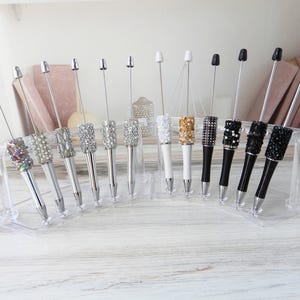May include: A collection of decorative pens with crystal and pearl embellishments. The pens have silver and black barrels, and are displayed on a clear acrylic stand. The pen tips are silver.