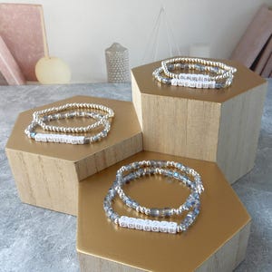 May include: Three silver bead bracelets with crystal accents on wooden display stands. The bracelets spell out the words "GRATEFUL", "GORGEOUS", and "DANIEL JASMINE".
