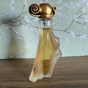 May include: A clear glass perfume bottle with a gold-colored cap and a unique, flowing design. The bottle contains a light amber liquid and is set against a floral patterned background.