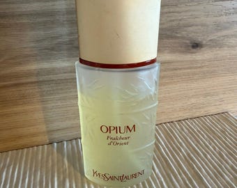 May include: A vintage perfume bottle of Opium by Yves Saint Laurent. The bottle has a frosted glass body with a light yellow liquid inside. The cap is a combination of beige and dark red. The text "Opium Fraicheur d'Orient" is printed on the bottle.