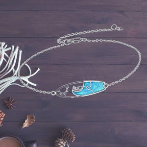 Op de afbeelding: Een zilveren ketting met een hanger in de vorm van een surfboard met een blauw opaal golfontwerp.