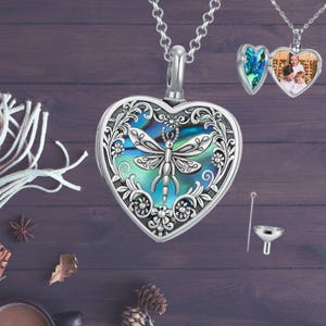 May include: Silver heart-shaped locket necklace with a dragonfly design and abalone shell inlay. The locket opens to reveal a small compartment for holding a photo or other small items. It comes with a funnel for filling the locket.
