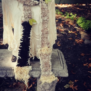 May include: A handmade scarf with a unique design. The scarf features sections of dark brown, beige, and cream-colored yarn, with a textured, fluffy appearance. Small green accents are incorporated throughout. The scarf is displayed on a mannequin.